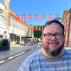 White man with short dark hair, glasses, and beard, in striped shirt, smiling on sunny street with red-and-white flags.