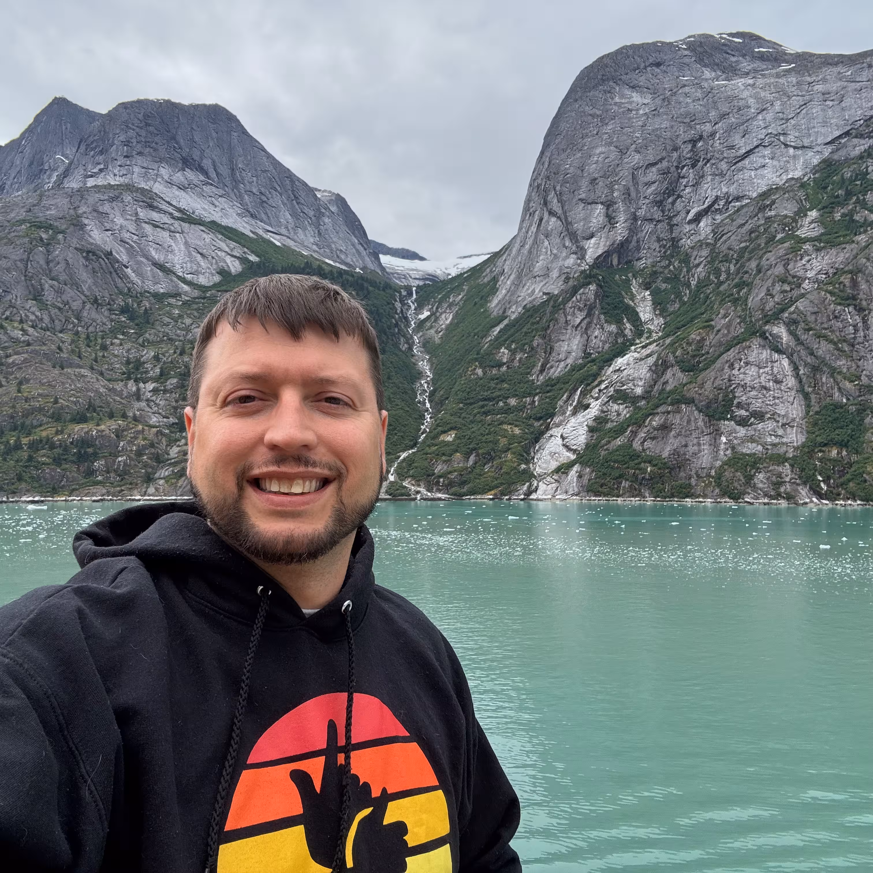 White man with light brown hair and beard smiling outdoors with greenery and mountains in the background.