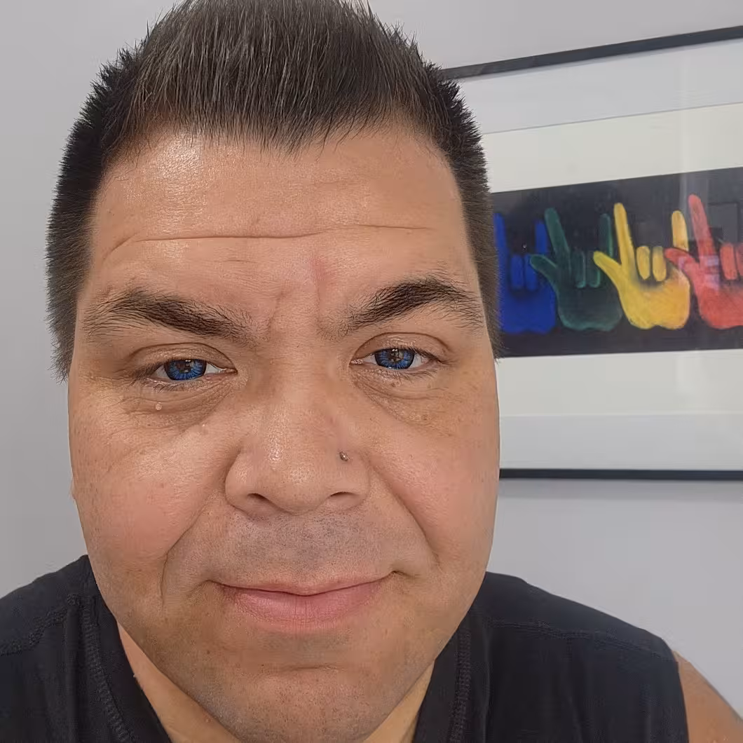 Native American man with short dark hair, blue eyes, nose piercing, in black shirt, smiling before artwork with colorful hand signs.