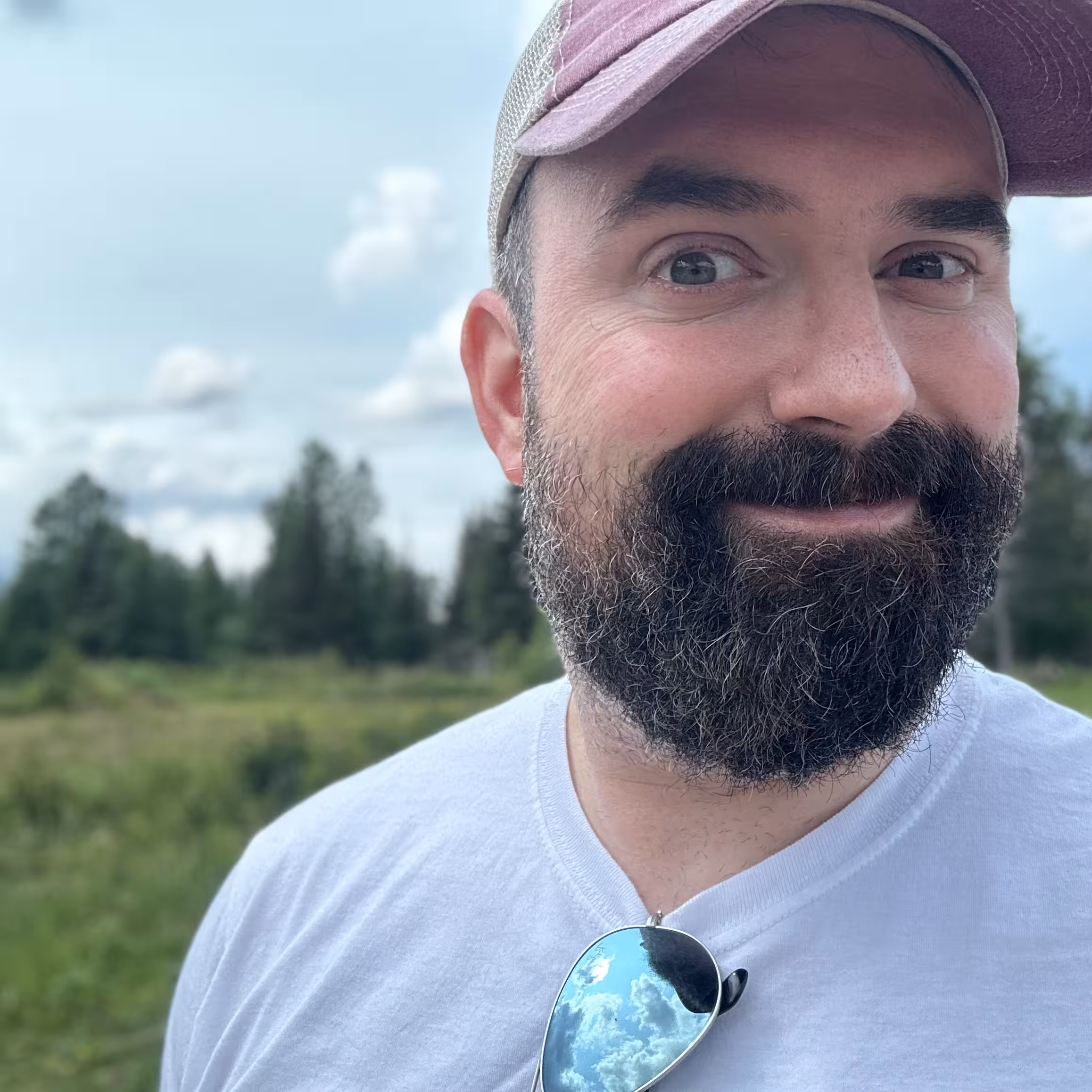 White man with full dark beard, cap, and white shirt with sunglasses clipped on, smiling outdoors in grassy landscape.